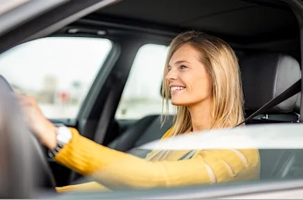Junge blonde Frau mit gelbem Pullover sitzt lächelnd am Steuer eines Autos.