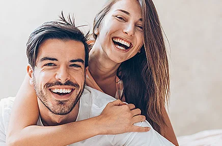 Glückliches Paar lacht zusammen im Bett, die Frau umarmt den Mann von hinten, beide tragen weiße Kleidung und strahlen Freude und Intimität aus.