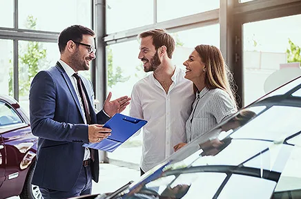 Ein Autoverkäufer im Anzug berät ein lächelndes Paar in einem hellen Autohaus mit mehreren Fahrzeugen im Showroom.