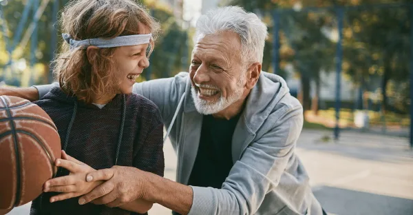 Ein älterer Mann mit grauem Bart und ein Kind mit Stirnband lachen gemeinsam beim Basketball spielen im Park an einem sonnigen Tag.