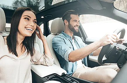 Ein lächelndes Paar sitzt im Auto, die Frau telefoniert am Handy, während der Mann fröhlich am Steuer fährt.