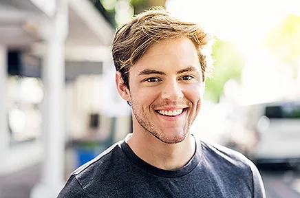 Ein junger Mann mit braunem Haar lächelt freundlich in die Kamera, er trägt ein dunkelblaues T-Shirt und steht in einer hellen, unscharfen Umgebung im Freien.