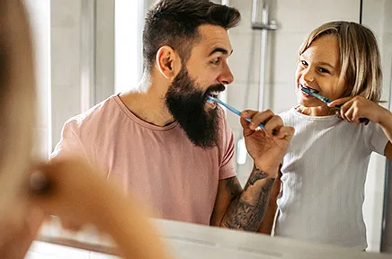 Vater und Tochter putzen gemeinsam lachend ihre Zähne vor dem Badezimmerspiegel und schauen sich dabei fröhlich an.