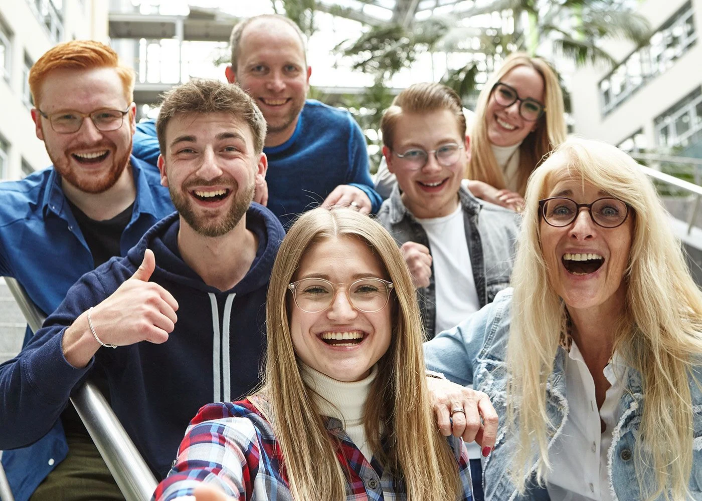 Eine fröhliche Gruppe von sieben Menschen unterschiedlichen Alters lächelt in die Kamera und macht ein Gruppenselfie in einem hellen Innenraum mit Glasdach.