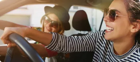 Zwei lächelnde Frauen mit Sonnenbrillen fahren gemeinsam im Auto, die Fahrerin trägt ein gestreiftes Shirt und beide genießen sichtlich die Fahrt bei Sonnenschein.