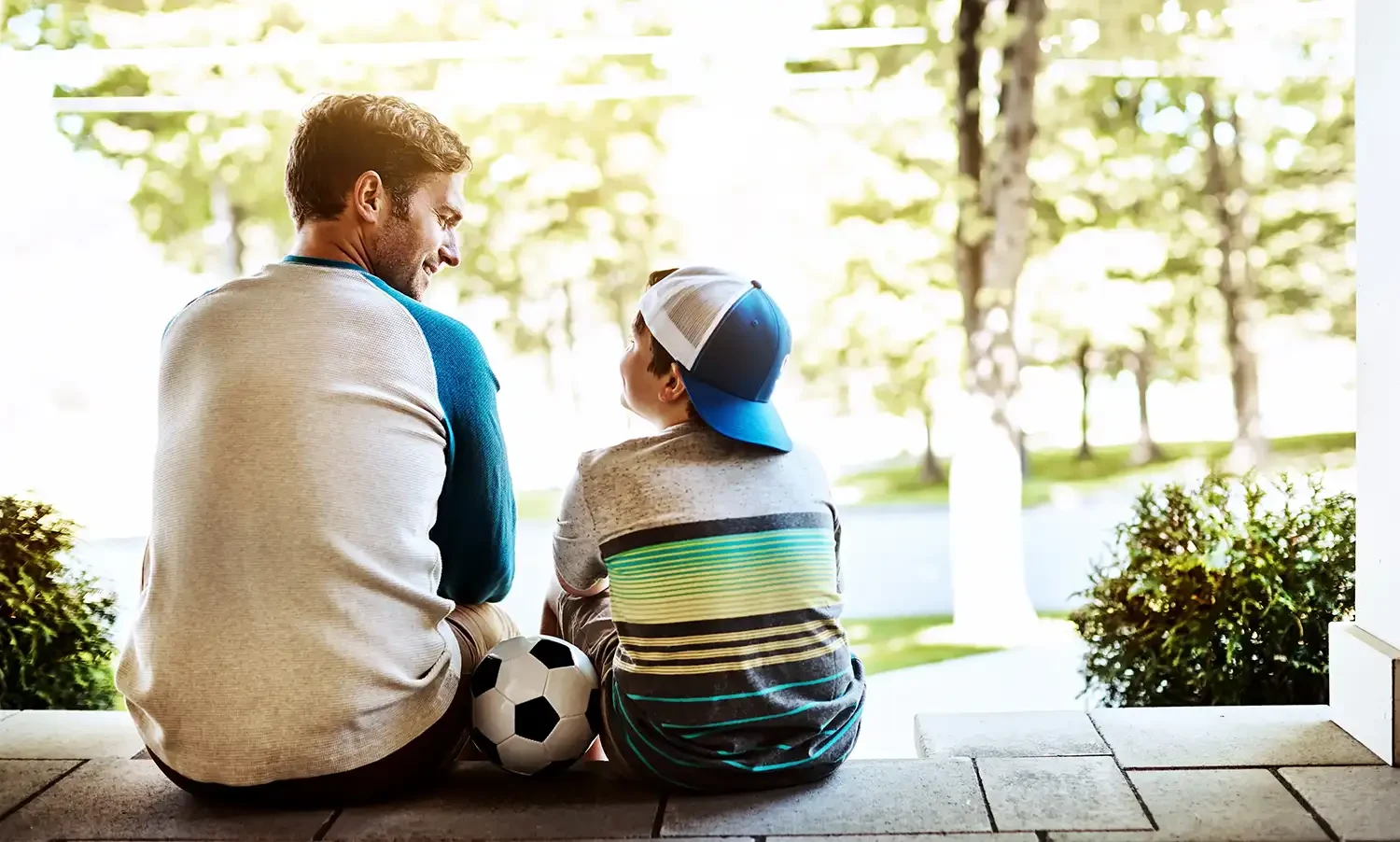 Ein Vater und sein Sohn sitzen Seite an Seite auf einer Terrasse, schauen gemeinsam in die Natur, ein Fußball liegt neben ihnen.