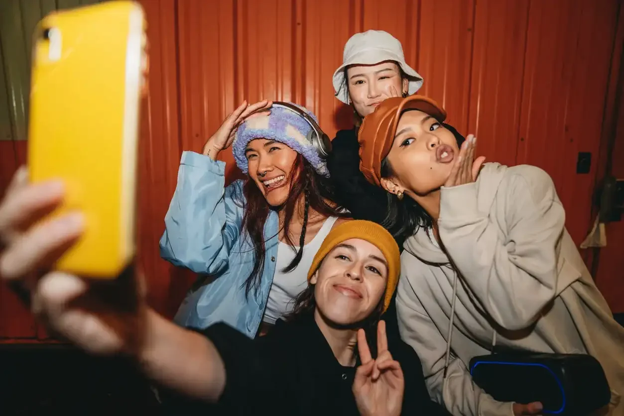 Vier junge Frauen mit bunten Mützen machen ein fröhliches Selfie vor einer roten Wellblechwand, eine macht ein Peace-Zeichen.