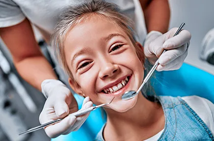 Ein fröhliches Kind sitzt im Zahnarztstuhl und lächelt in die Kamera, während eine Zahnärztin mit Handschuhen und Instrumenten eine Untersuchung durchführt.