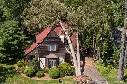 Luftaufnahme eines Fachwerkhauses mit rotem Dach, umgeben von grünen Bäumen und gepflegtem Garten mit runden Büschen und einer Auffahrt.