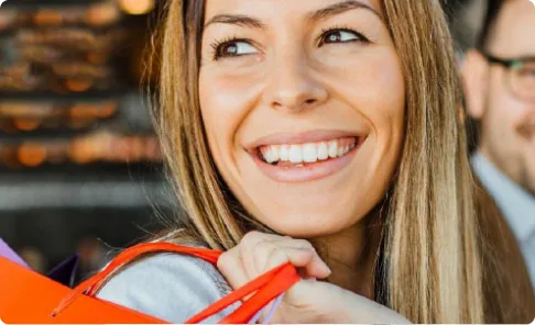 Lächelnde Frau mit langen blonden Haaren trägt eine orange Tasche über der Schulter, im Hintergrund sind weitere Personen unscharf zu sehen.