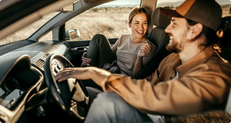 Ein lächelndes Paar sitzt im Auto, die Frau auf dem Beifahrersitz lacht den Mann am Steuer an, während sie gemeinsam eine entspannte Autofahrt genießen.