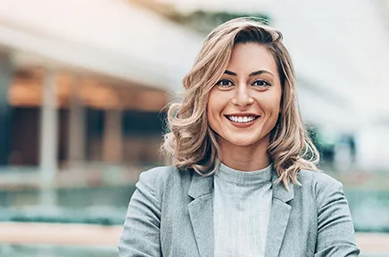 Lächelnde Geschäftsfrau mit blonden Haaren in grauem Blazer steht mit verschränkten Armen vor modernem Bürogebäude.