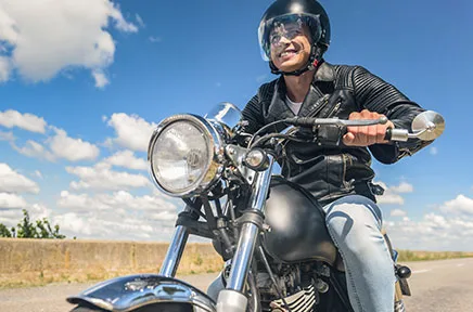 Motorradfahrer in schwarzer Lederjacke und Helm fährt auf einer Landstraße unter blauem Himmel mit weißen Wolken.