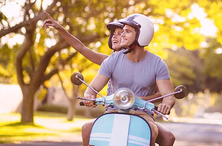 Junges Paar fährt fröhlich auf einem hellblauen Motorroller durch einen sonnigen Park mit herbstlichem Laubwerk, beide tragen Helme.