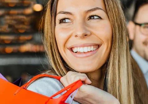 Lächelnde Frau mit langen blonden Haaren hält eine orange-rote Tasche und blickt fröhlich zur Seite in einem Geschäft oder Café.