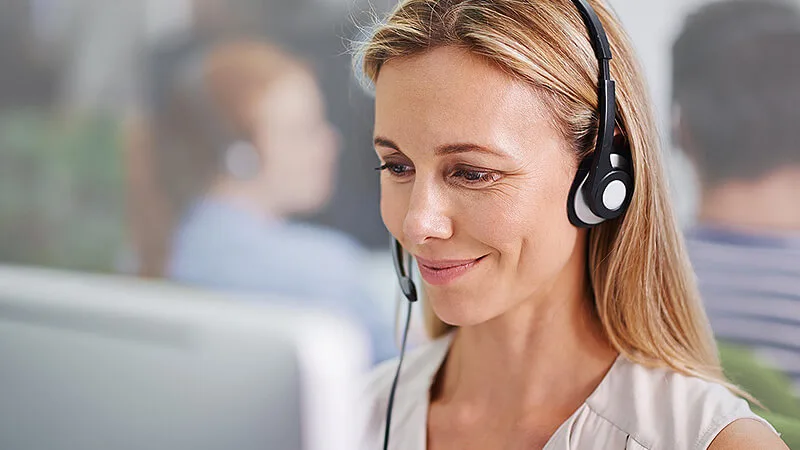 Eine lächelnde Frau mit Headset arbeitet an einem Computer in einem modernen Büro, vermutlich im Kundenservice oder Support.
