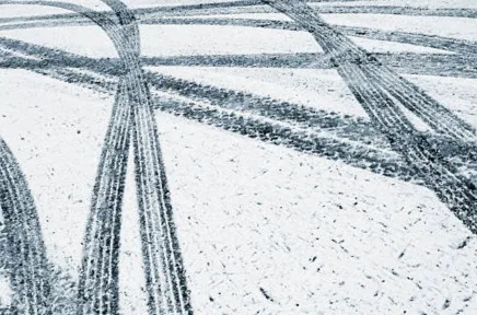 Reifenspuren von mehreren Fahrzeugen kreuzen sich auf einer schneebedeckten Straße oder einem verschneiten Platz und bilden ein Muster aus dunklen Linien im weißen Schnee.