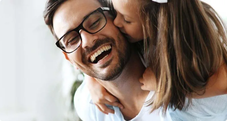 ein kind umarmt von hinten einen lächelnden mann mit brille, der sie huckepack trägt, in einer liebevollen und fröhlichen geste.