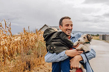 Lächelnder Mann in Jeansjacke hält kleinen Hund vor einem Maisfeld unter bewölktem Himmel.