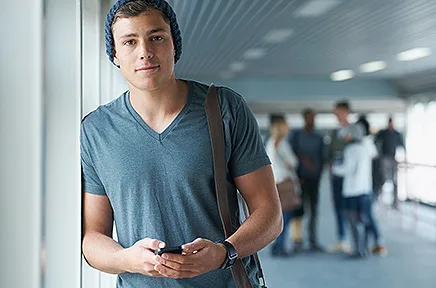 Junger Mann mit Rucksack und Smartphone steht lächelnd in einem modernen Gebäudeflur, während im Hintergrund weitere Personen zu sehen sind.