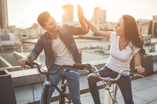 Ein junges Paar auf Fahrrädern gibt sich ein High-Five auf einer Dachterrasse mit Stadtpanorama im Hintergrund bei Sonnenschein.