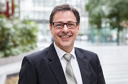 Lächelnder Geschäftsmann mit Brille in dunklem Anzug und heller Krawatte steht in einer urbanen Umgebung mit unscharfem Hintergrund aus Gebäuden und Grünflächen.