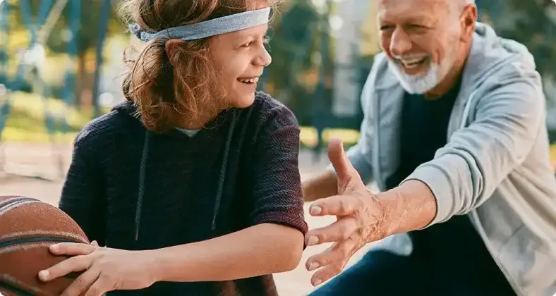 Ein älterer Mann und ein Kind mit Stirnband spielen gemeinsam Basketball im Park und haben dabei sichtlich Spaß.