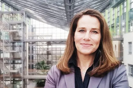 Frau mit schulterlangem braunem Haar in grauer Jacke und schwarzem Oberteil steht in einem modernen Gebäude mit Glasarchitektur und Atrium im Hintergrund.