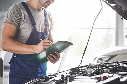 Ein Kfz-Mechaniker in blauer Arbeitslatzhose notiert Informationen auf einem Klemmbrett, während er vor einem geöffneten Motorraum eines Fahrzeugs in einer hellen Werkstatt steht.