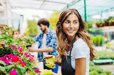 Lächelnde Frau mit langen braunen Haaren in Schürze hält Blumentopf in einem Gartencenter, im Hintergrund arbeitet ein Mann zwischen bunten Pflanzen.